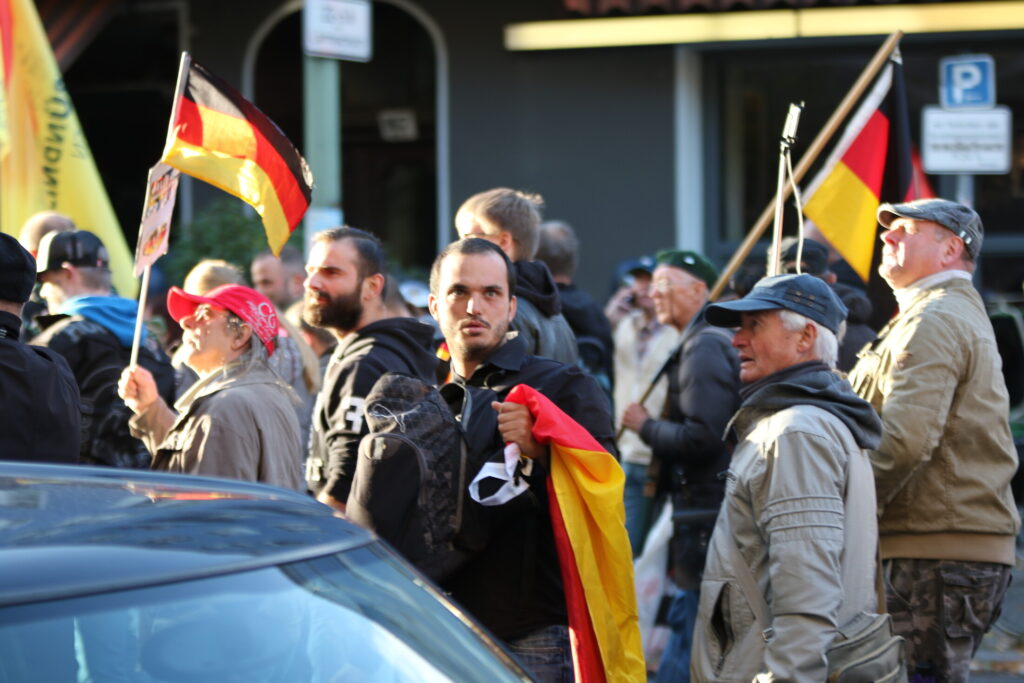 Alexander Scheuermann steht in der Bildmitte und schaut leicht nach oben, er trägt einen dunklen Pullover und eine helle Jeans. In der linken Hand hält er eine Deutschland Fahne, seinen Rucksack trägt er vorne. Um ihn herum sind weitere Teilnehmende des Aufmarsches sowie Deutschlandfahnen zu sehen.